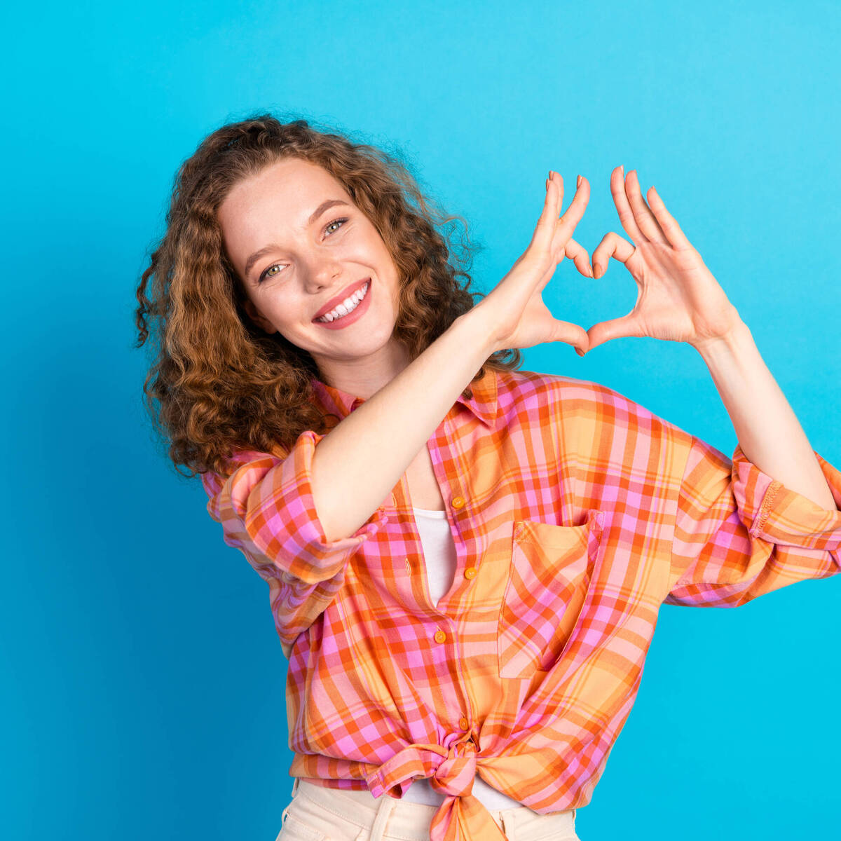 Joyful young woman with red hair makes heart gesture, showcasing happiness on a vibrant blue background